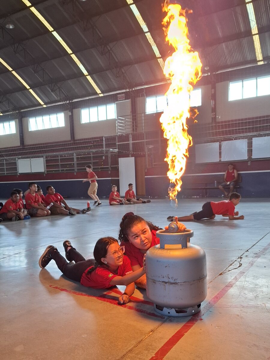 Aluna e instrutora deitadas no chão do ginásio durante treinamento de prevenção com botijão de gás e chama controlada