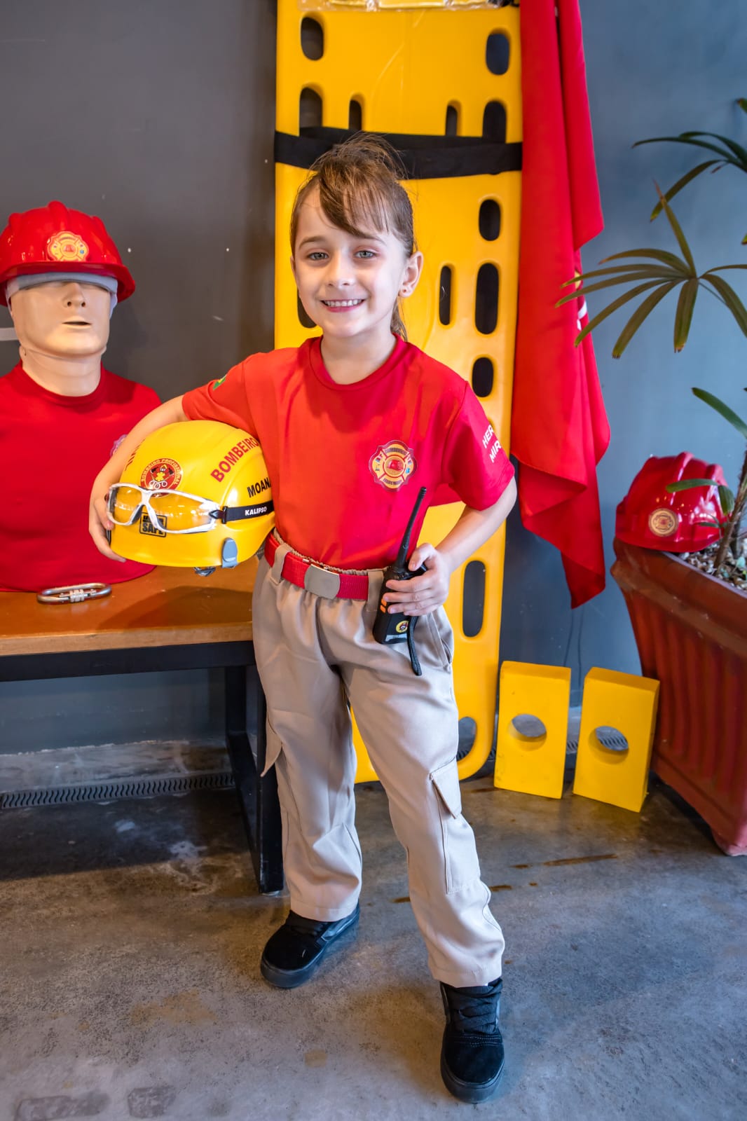 Bombeira mirim sorrindo com capacete e rádio, ao lado da maca de resgate